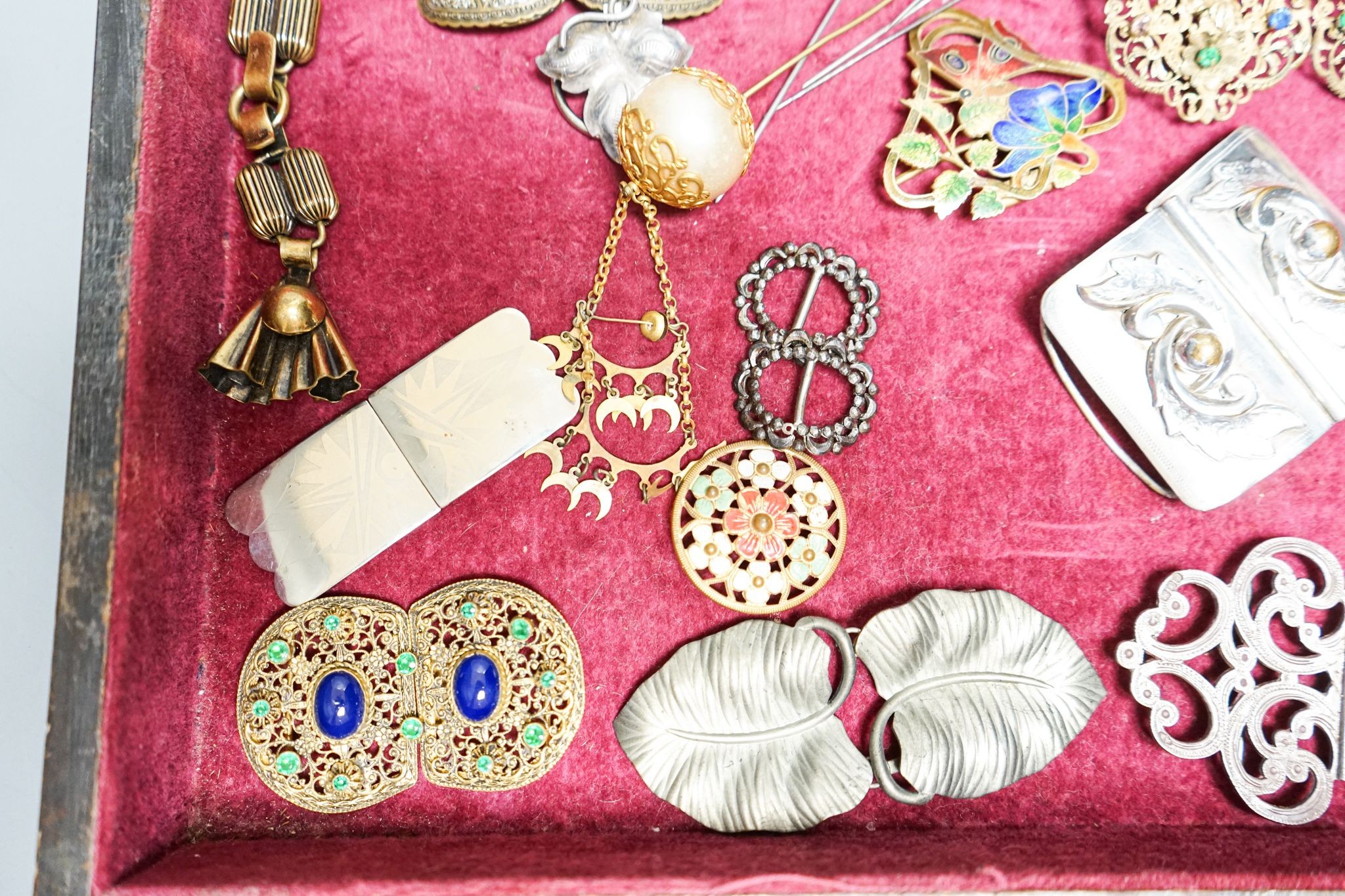 A group of belt buckles, clips and hatpins in a mahogany display case, 30.5cm